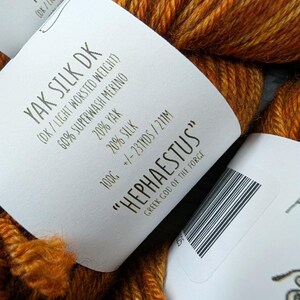 May include: Close-up of a skein of orange and brown Yak Silk DK yarn. The label reads "HEPHAESTUS" and "Greek God of the Forge". The yarn is a blend of Yak, Superwash Merino, and Silk. The label also indicates the weight and yardage.