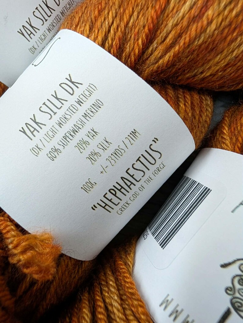 May include: Close-up of a skein of orange and brown Yak Silk DK yarn. The label reads "HEPHAESTUS" and "Greek God of the Forge". The yarn is a blend of Yak, Superwash Merino, and Silk. The label also indicates the weight and yardage.