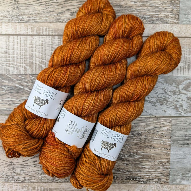 May include: Three skeins of orange-brown yarn with white labels that read "ATHC WOOLS" and "Yak Silk DK". The yarn is a rich, warm color, perfect for knitting or crocheting projects. The labels also feature a sheep logo and the text "HERPHAESTUS".