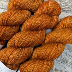 May include: Close-up of several skeins of orange yarn. The yarn is a rich, warm orange color with subtle variations in tone. The texture of the yarn is visible, suggesting a soft and luxurious material. The yarn is labeled "HEPHAESTUS".