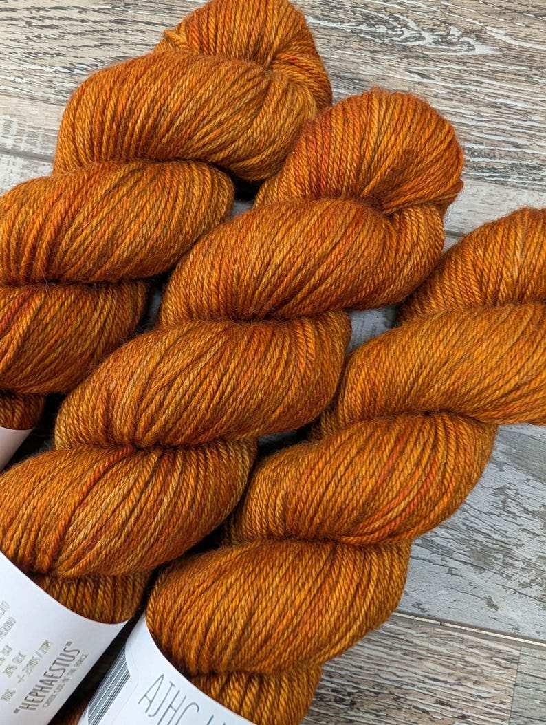 May include: Close-up of several skeins of orange yarn. The yarn is a rich, warm orange color with subtle variations in tone. The texture of the yarn is visible, suggesting a soft and luxurious material. The yarn is labeled "HEPHAESTUS".