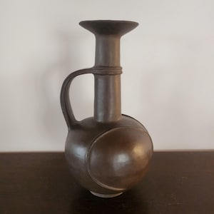 Puede incluir: Un jarrón de cerámica marrón con cuerpo redondeado y cuello largo. El jarrón tiene un asa y un diseño circular decorativo en el lateral. El jarrón mide aproximadamente 25 cm de altura.