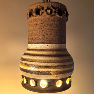 Pode incluir: Luminária pendente de cerâmica vintage. A lâmpada tem formato cilíndrico com uma seção superior marrom texturizada e um padrão listrado em tons de marrom, branco e dourado. A parte inferior apresenta recortes circulares. A luz está acesa.