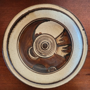 May include: A round, ceramic plate with a cream-colored rim and a brown, abstract design in the center. The design features concentric circles and curved shapes in shades of brown and tan. The plate has a rustic, handmade appearance.