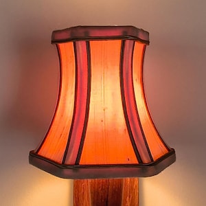 Puede incluir: Una pantalla de lámpara vintage con una forma y diseño únicos. La pantalla presenta un tono naranja cálido con rayas rojas verticales y un borde marrón oscuro. Está montada sobre una base de madera, creando una estética clásica.