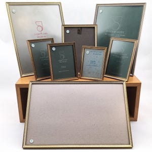 Può includere: Una collezione di cornici dorate di varie dimensioni, alcune con retro verde o grigio. Le cornici hanno un bordo decorativo con perline. Alcune cornici riportano la scritta "JYDEN QUALITY" e "ANTI REFLEX GLAS 8 x 10,5 cm".