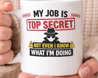 May include: White ceramic mug with the text "MY JOB IS TOP SECRET" in black and red. Below the text is a graphic of a person in a hat and sunglasses. The mug also says "NOT EVEN I KNOW WHAT I'M DOING."