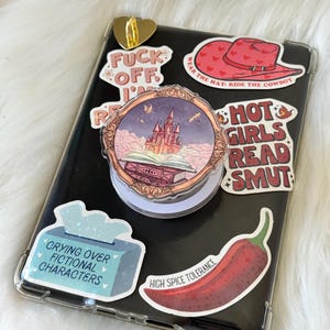 May include: A black phone in a clear case is decorated with stickers. Stickers include a red cowboy hat, a castle scene, a tissue box that says "Crying Over Fictional Characters", and a red chili pepper. A gold heart pin is at the top.