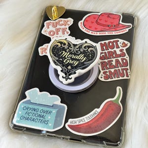 Puede incluir: Una funda transparente para teléfono adornada con varias pegatinas. Las pegatinas incluyen frases como "Fuck Off", "Morally Grey" y "Hot Girls Read Smut". Otras pegatinas presentan un sombrero de vaquero rojo, un chile rojo y una caja de pañuelos.