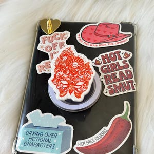 Könnte beinhalten: Eine Sammlung von Aufklebern auf einer schwarzen Handyhülle. Zu den Aufklebern gehören ein roter Cowboyhut, eine rote Chilischote und Texte wie "FUCK OFF", "HOT GIRLS READ SMUT" und "CRYING OVER FICTIONAL CHARACTERS". Ein goldener herzförmiger Handyhalter ist ebenfalls sichtbar.