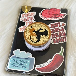 May include: A black smartphone in a clear case, adorned with stickers. Stickers feature a red cowboy hat, a chilli pepper, and text saying "Hot Girls Read Smut". A pop socket with a dragon design is attached.