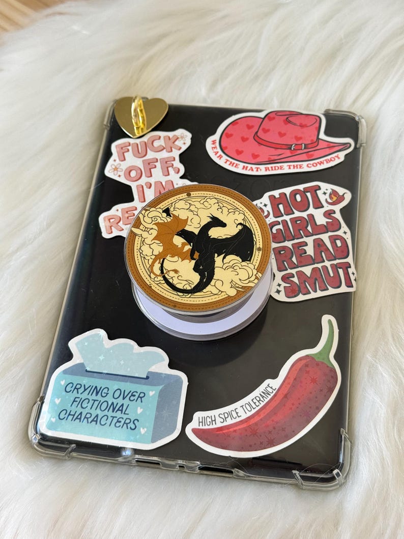 K&ouml;nnte beinhalten: Ein schwarzes Smartphone mit einer transparenten H&uuml;lle ist mit Aufklebern versehen. Zu den Aufklebern geh&ouml;ren ein roter Cowboyhut, eine Chilischote und Text mit der Aufschrift "Hot Girls Read Smut". Ein Pop-Socket mit Drachendesign ist angebracht.