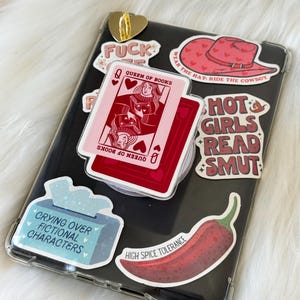 May include: A phone case with multiple stickers. Stickers include a red cowboy hat, a red chili pepper, a playing card that says "Queen of Books", and text that says "Hot Girls Read Smut".
