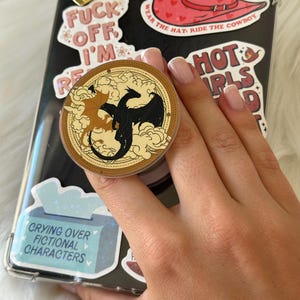 K&ouml;nnte beinhalten: Eine schwarze Handyh&uuml;lle mit verschiedenen Aufklebern, darunter ein roter Cowboyhut, ein goldenes Herz und Text mit der Aufschrift "FUCK OFF I'M". Ein runder Pop-Socket zeigt zwei Drachen. Der Text "CRYING OVER FICTIONAL CHARACTERS" ist ebenfalls sichtbar.