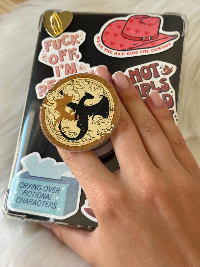 K&ouml;nnte beinhalten: Eine schwarze Handyh&uuml;lle mit verschiedenen Aufklebern, darunter ein roter Cowboyhut, ein goldenes Herz und Text mit der Aufschrift "FUCK OFF I'M". Ein runder Pop-Socket zeigt zwei Drachen. Der Text "CRYING OVER FICTIONAL CHARACTERS" ist ebenfalls sichtbar.