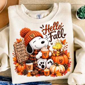 May include: A cream-colored crewneck sweatshirt featuring a graphic of a white dog wearing an orange knit hat and plaid scarf, holding an orange mug. Text reads Hello Fall and Sweater Weather Pumpkin Spice & Everything Nice with pumpkins.