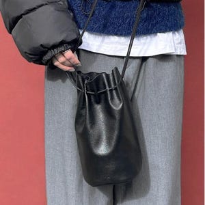 Puede incluir: Un bolso tipo cubo de cuero negro con cierre de cord&oacute;n y una correa larga y fina. El bolso lo sostiene una persona que lleva una chaqueta acolchada negra, un su&eacute;ter azul y pantalones grises. El fondo es rojo liso.