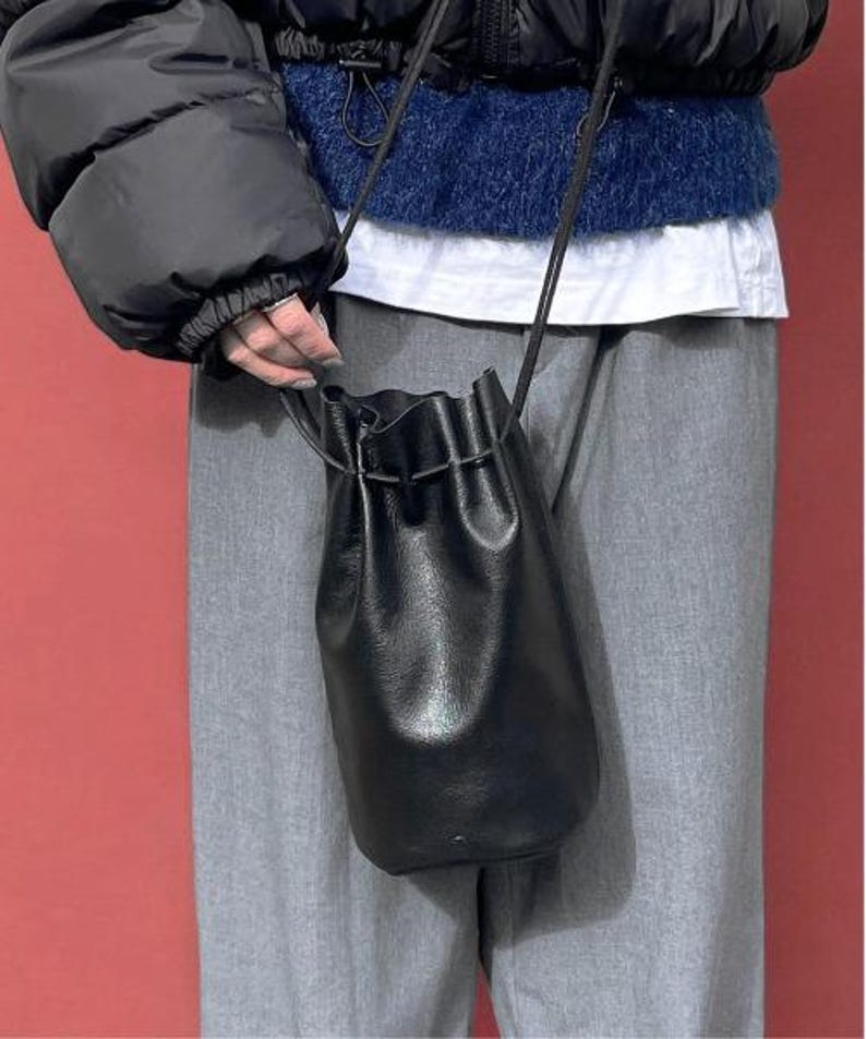 Puede incluir: Un bolso tipo cubo de cuero negro con cierre de cord&oacute;n y una correa larga y fina. El bolso lo sostiene una persona que lleva una chaqueta acolchada negra, un su&eacute;ter azul y pantalones grises. El fondo es rojo liso.