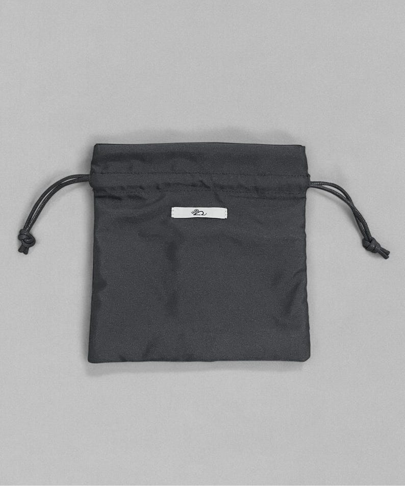 Puede incluir: Una bolsa negra con cord&oacute;n y una peque&ntilde;a etiqueta blanca. La bolsa est&aacute; hecha de una tela lisa y tiene forma rectangular. El cord&oacute;n es negro y est&aacute; anudado en ambos lados. La etiqueta tiene un peque&ntilde;o dise&ntilde;o estilizado.
