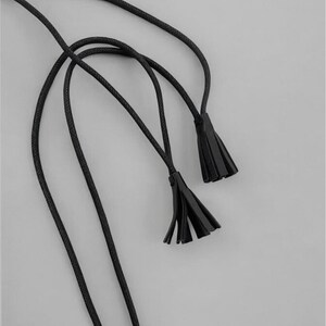 Puede incluir: Primer plano de una cuerda negra con dos borlas negras sobre un fondo gris. La cuerda est&aacute; finamente trenzada y las borlas est&aacute;n hechas de un material similar. El dise&ntilde;o es simple y elegante, adecuado para varios proyectos de artesan&iacute;a o accesorios.