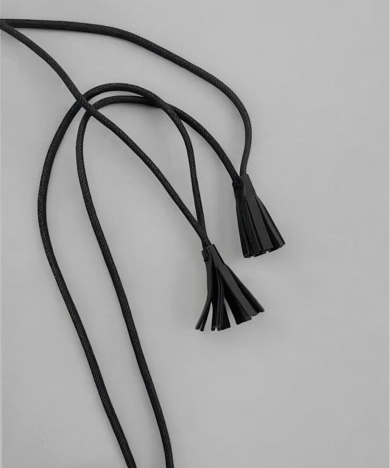 Puede incluir: Primer plano de una cuerda negra con dos borlas negras sobre un fondo gris. La cuerda est&aacute; finamente trenzada y las borlas est&aacute;n hechas de un material similar. El dise&ntilde;o es simple y elegante, adecuado para varios proyectos de artesan&iacute;a o accesorios.