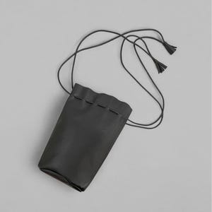 Puede incluir: Una bolsa de cuero negro con cierre de cord&oacute;n y una correa larga y fina. La bolsa tiene forma rectangular con un borde superior ligeramente ondulado. La correa termina en peque&ntilde;as borlas. El fondo es gris liso.