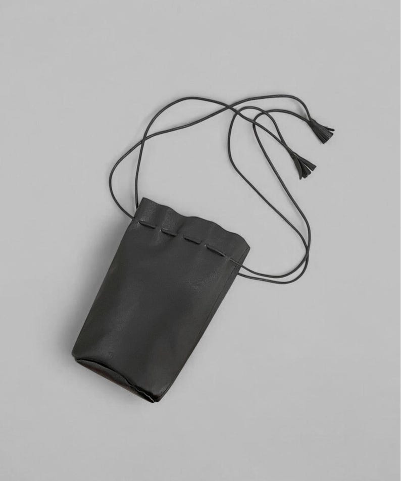Puede incluir: Una bolsa de cuero negro con cierre de cord&oacute;n y una correa larga y fina. La bolsa tiene forma rectangular con un borde superior ligeramente ondulado. La correa termina en peque&ntilde;as borlas. El fondo es gris liso.
