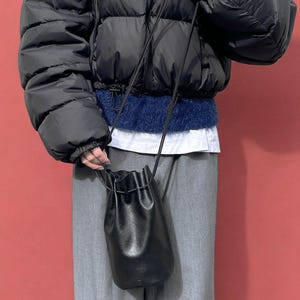 Puede incluir: Un bolso tipo cubo negro de cuero con cord&oacute;n y una correa larga y fina. El bolso lo sostiene una persona que lleva una chaqueta acolchada negra, un su&eacute;ter azul y pantalones grises. El fondo es de color rojo liso.