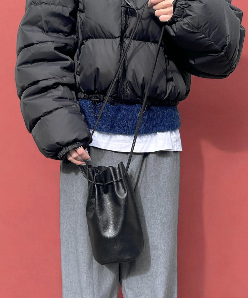 Puede incluir: Un bolso tipo cubo negro de cuero con cord&oacute;n y una correa larga y fina. El bolso lo sostiene una persona que lleva una chaqueta acolchada negra, un su&eacute;ter azul y pantalones grises. El fondo es de color rojo liso.