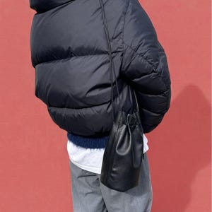 Puede incluir: Un bolso de cubo de cuero negro con cierre de cord&oacute;n cuelga de una fina correa. El bolso se muestra sobre un fondo rojo, usado con una chaqueta acolchada azul marino y pantalones grises.