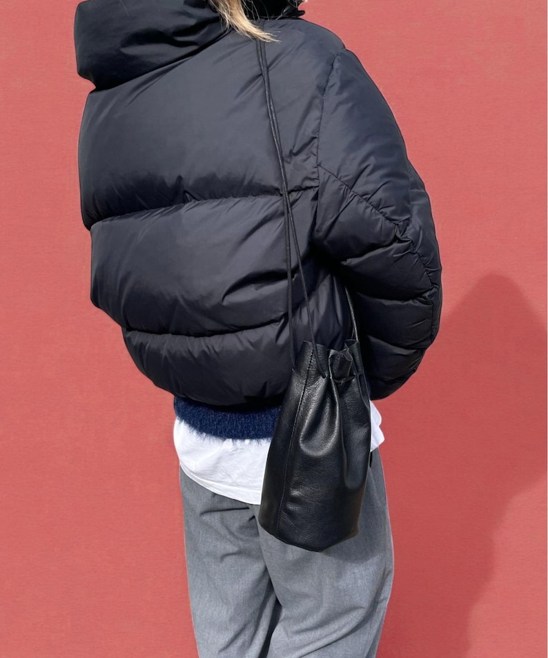 Puede incluir: Un bolso de cubo de cuero negro con cierre de cord&oacute;n cuelga de una fina correa. El bolso se muestra sobre un fondo rojo, usado con una chaqueta acolchada azul marino y pantalones grises.