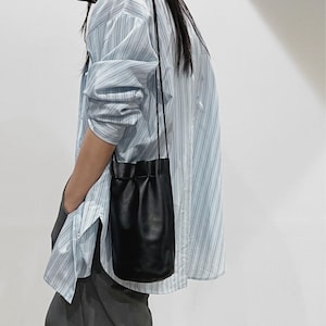 Puede incluir: Un bolso tipo cubo de cuero negro con cierre de cord&oacute;n cuelga de una fina correa. El bolso se lleva sobre una camisa abotonada a rayas azul claro y blanco y pantalones grises oscuros.