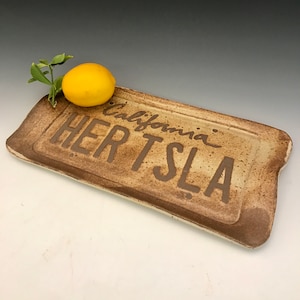 Könnte beinhalten: Ein Keramik-Serviertablett mit brauner Glasur und weißem Hintergrund. Das Tablett trägt die Aufschrift "California" und "Hertsela" in braunen Lettern. Ein gelber Zitron sitzt auf dem Tablett.