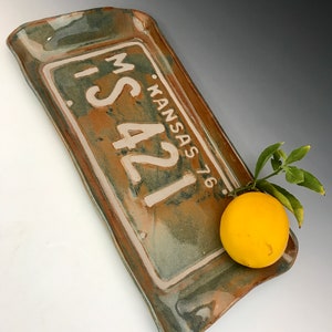 Könnte beinhalten: Ein Keramik-Serviertablett mit grüner und brauner Glasur. Das Tablett ist in Form eines Nummernschilds mit dem Text "M. KANSAS 76. IS421" in Weiß. Eine gelbe Zitrone liegt auf dem Tablett.