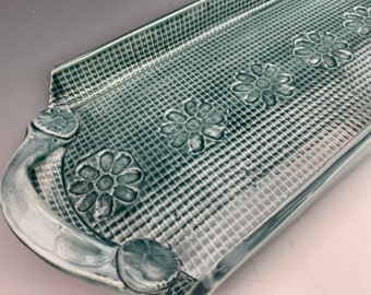May include: A rectangular serving tray with two handles, made of glazed ceramic. The tray has a green and white checkered pattern with raised flower designs.