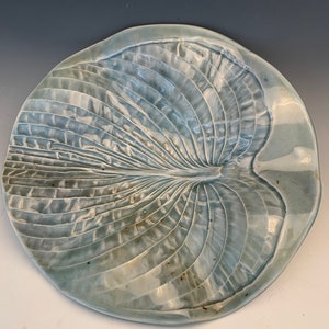 May include: A light blue ceramic plate with a raised design of a leaf. The plate has a slightly irregular shape and a glossy finish.