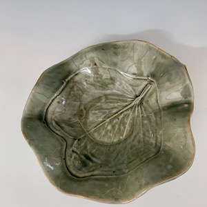 May include: A green ceramic bowl shaped like a leaf. The bowl has a textured surface and a raised leaf design in the center.