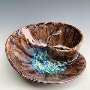 May include: A ceramic serving dish with a spiral design, featuring a glossy glaze in shades of deep brown, tan, and a vibrant turquoise blue center. The bowl has a curved, shell-like shape with a raised, integrated circular compartment.