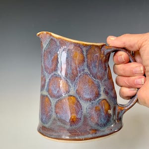 Może przedstawiać: Brązowy dzbanek ceramiczny z niebiesko-białym szkliwem. Dzbanek ma zaokrąglone ciało i uchwyt. Szkliwo ma fakturowaną, bąbelkową powierzchnię.