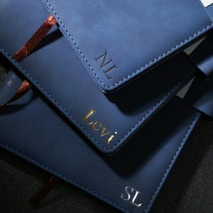 Może przedstawiać: Zbliżenie na trzy granatowe skórzane notatniki. Górny notatnik ma inicjały "NL" wytłoczone w srebrze. Środkowy notatnik ma imię "Levi" wytłoczone w złocie. Dolny notatnik ma inicjały "SL" wytłoczone w srebrze. Każdy notatnik ma szytą obwódkę.