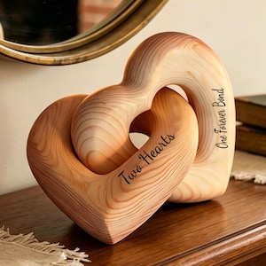 Peut inclure: Une sculpture en bois clair composée de deux cœurs entrelacés. Le grain du bois est visible sur toute la pièce. Le texte "Two Hearts" est gravé sur le cœur avant, et le texte "One Forever Bond" est gravé sur le côté de la sculpture.