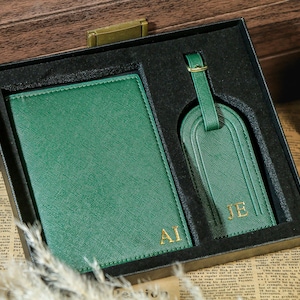 Könnte beinhalten: Ein grünes Reisepass- und Gepäckanhänger-Set mit goldenen Initialen in einer schwarzen Box. Der Reisepasshalter und der Gepäckanhänger bestehen aus einem strukturierten Material. Die Initialen "AI" und "JE" sind golden.