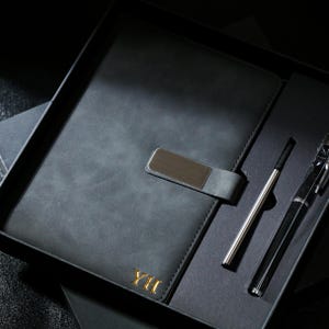 May include: A black leather journal with a silver clasp and gold lettering, presented in a gift box. Includes a silver pen and a black pen. The journal measures approximately 8 x 6 inches.