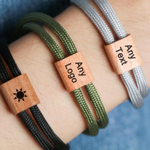 Può includere: Primo piano di tre braccialetti in corda intrecciata neri, verde oliva e grigi. Ogni braccialetto presenta una perla di legno con il testo "Any Text" o "Any Logo" e un motivo a sole. I braccialetti sono indossati su un polso.