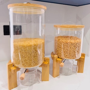 Könnte beinhalten: Zwei transparente Glas-Vorratsbehälter mit Bambusdeckeln und Holzständern. Die Behälter sind mit Getreide gefüllt und haben transparente Messbecher. Auf einer weißen Arbeitsplatte.