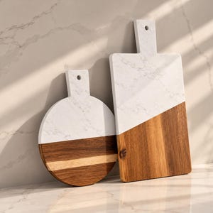 May include: Two serving boards featuring a combination of white marble with grey veining and natural brown wood. One board is circular and the other is rectangular, both equipped with a handle containing a small hole for hanging.