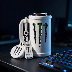 Może przedstawiać: Biały cylindryczny uchwyt na puszkę z rączką, posiadający wycięcie z zielono-czarnym logo Monster Energy. Do rączki przymocowana jest biała okrągła podstawka oraz mała biała prostokątna zawieszka, obie z widocznym logo Monster Energy.