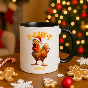 Puede incluir: Taza de cerámica blanca con interior y asa negros. La taza presenta un dibujo de un pollo de dibujos animados con gorro de Papá Noel y luces navideñas, con el texto "F-CAW-F" y el nombre "JACKIE". Tema festivo navideño.