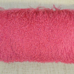 May include: A pink fluffy pillow with long, curly fibers. The pillow is made of a soft, plush material and has a rectangular shape.