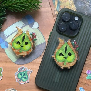 Op de afbeelding: Een groene telefoonhoes met een telefoonhouder van een cartoonkarakter bevestigd. De grip heeft een bladachtig ontwerp met een gezicht en kleine rode bessen. Verschillende stickers van hetzelfde karakter en andere ontwerpen zijn verspreid over een houten oppervlak.